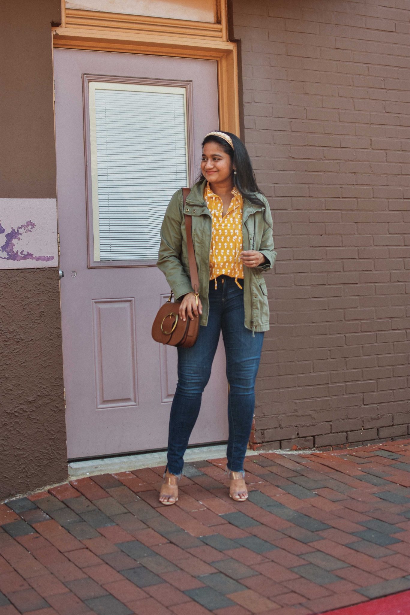 Wearing J.Crew Ruffle Sleeve Cotton voile top, AG Farrah ankle skinny jeans, Schutz Ariella mule, Madewell utlity Passage Jacket | Lightweight Jackets by popular Maryland modest fashion blog, Dreaming Loud: image of a woman wearing a utility jacket, yellow and white blouse, jeans, gold strap sandals, cream colored headband, and brown purse. 